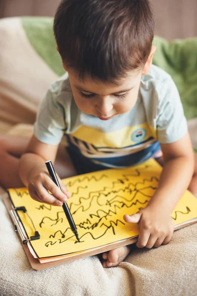 Caucásico niño pequeño está dibujando en un papel amarillo usando un marcador mientras sititng en la cama en el día de los niños —  Fotos de Stock