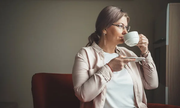 Foto vista lateral de una mujer mayor bebiendo una taza de té mientras mira a través de la ventana desde el interior de la casa — Foto de Stock
