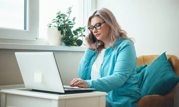 Caucasian senior businesswoman with glasses and blonde hair is talking on phone while working remotely with a laptop — Stock Photo, Image