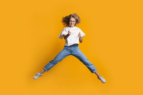 Salto donna bionda sta gesticolando il colpo e pistola con entrambe le mani mentre salta su un muro giallo in studio — Foto Stock