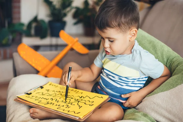 Precioso chico morena dibujando en un papel amarillo usando un marcador mientras está sentado en almohadas en casa durante el día internacional de los niños —  Fotos de Stock