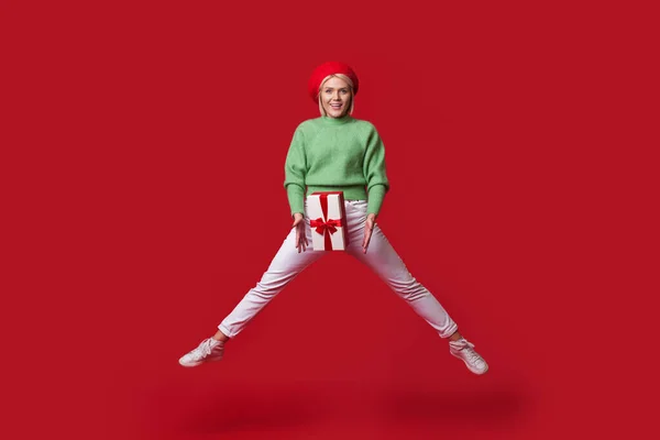 Блондинка модна жінка стрибає на червоній стіні тримає подарункову коробку і носить капелюх, посміхаючись на камеру — стокове фото