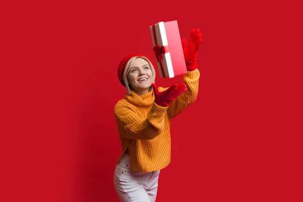 Uśmiechnięta biała kobieta nosi rękawiczki i kapelusz na czerwonej ścianie podczas łapania pudełka prezentów — Zdjęcie stockowe