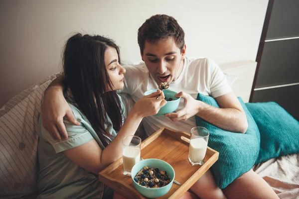 Bruna donna e il suo amante sono seduti a letto che si nutrono a vicenda con cereali e latte durante una mattina passata insieme — Foto Stock