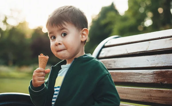 Rapaz bonito está comendo sorvete e olhando surpreso para a câmera enquanto sentado no banco no parque — Fotografia de Stock