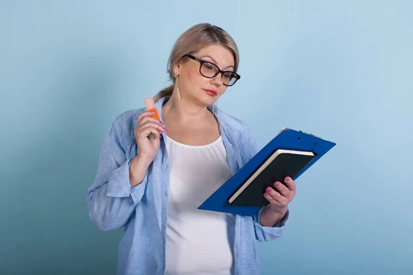Starsza kaukaska kobieta czyta coś podczas pozowania na niebieskiej ścianie w okularach i trzyma kilka folderów — Zdjęcie stockowe