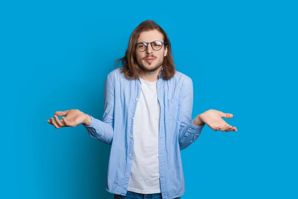 Homem caucasiano confuso com barba e cabelos longos está posando indeciso enquanto gesticula com palmas em uma parede de estúdio azul — Fotografia de Stock