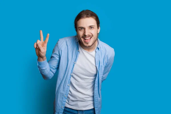Barbudo hombre caucásico está haciendo un gesto de la señal de hola y la paz sonriendo a la cámara en una pared azul en ropa casual — Foto de Stock