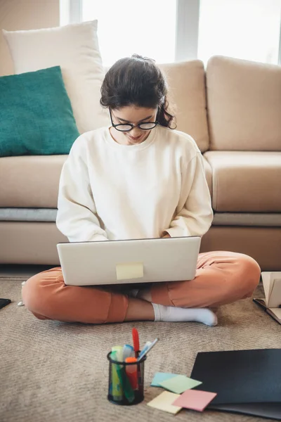 Gündelik giysiler içinde hoş bir bayan yerde oturuyor ve ödevlerini yaparken dizüstü bilgisayar kullanıyor ve gülümsüyor. — Stok fotoğraf