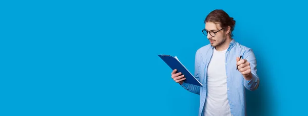 Jovem empresário está posando em uma parede azul com espaço livre olhando em uma pasta e segurando uma caneta — Fotografia de Stock