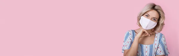 Fehér nő szőke haj visel nyári ruha mosolyog a kamera egy orvosi maszk pózol egy rózsaszín fal szabad hely — Stock Fotó