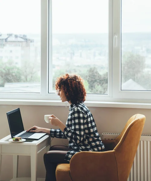 Kıvırcık saçlı, günlük kıyafetler giymiş beyaz kadın laptopta çalışırken sandviçle kahve içiyor. — Stok fotoğraf