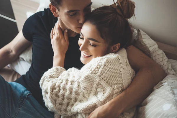 Καυκάσιος άντρας φιλάει τη χαριτωμένη γυναίκα του ντυμένη με ένα λευκό πλεκτό πουλόβερ που αγκαλιάζεται στο κρεβάτι — Φωτογραφία Αρχείου