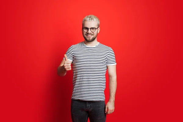 Homem caucasiano com cabelo loiro e óculos está gesticulando o sinal semelhante sorrindo para a câmera em uma parede de estúdio vermelha — Fotografia de Stock