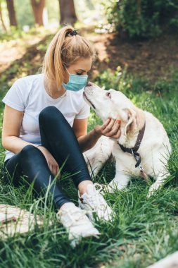 Maskeli sarışın beyaz kadın dışarıda köpeğiyle parkta çimlerin üzerinde eğleniyor.