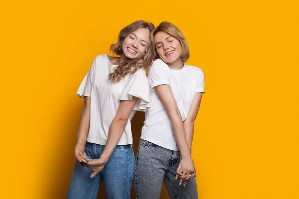 Sœurs blondes posent ensemble sur un mur jaune gesticulant bonheur avec les yeux fermés — Photo