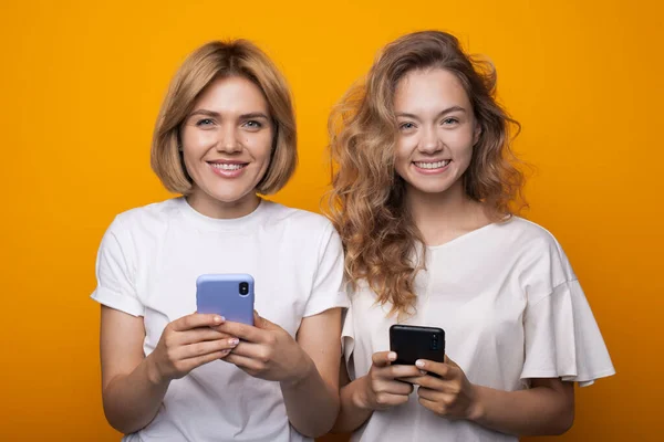 Två kaukasiska blonda systrar chattar med telefoner medan de ler mot kameran på en gul studiovägg — Stockfoto