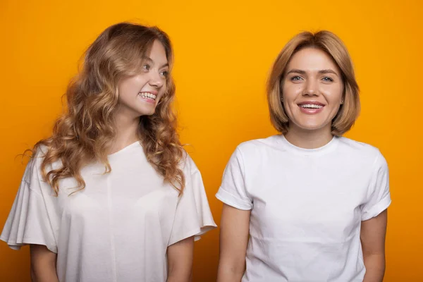 Donna caucasica con i capelli biondi che guarda sorridere a sua sorella in t-shirt bianca su un muro di studio giallo — Foto Stock