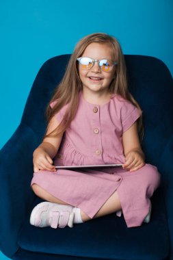 Gözlüklü tatlı sarışın kız mavi bir stüdyo duvarındaki koltukta tabletle oynuyor ve gülümsüyor.