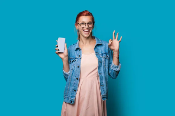 Mujer de jengibre caucásico está haciendo un gesto de la señal de bien mientras que muestra la pantalla del teléfono con espacio vacío en la cámara posando en la pared del estudio azul — Foto de Stock
