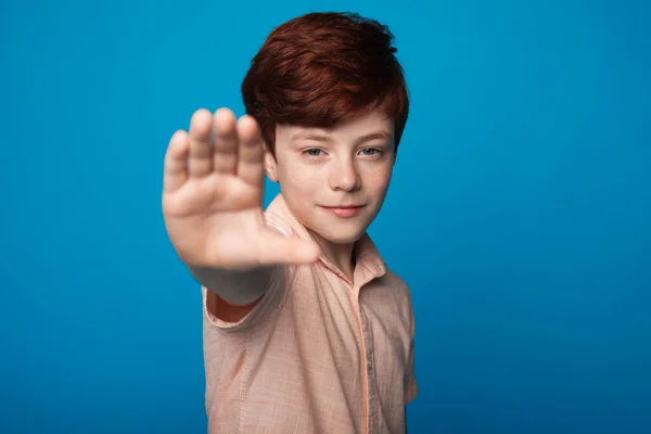 Feche a foto de um menino sorridente com cabelo vermelho e sardas gesticulando sinal de parada em uma parede azul olhando para a câmera — Fotografia de Stock