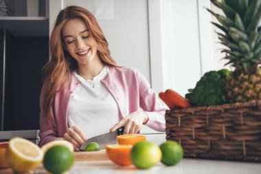 Çilli, kızıl saçlı, sevimli beyaz kadın bıçakla limon dilimliyor. Meyveli sepetin yanında salata yapıyor.