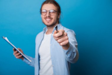 Gözlüklü ve uzun saçlı genç sakallı işadamı elinde bazı belgeler tutuyor ve kameraya dişleriyle gülümseyen kalemle işaret ediyor.