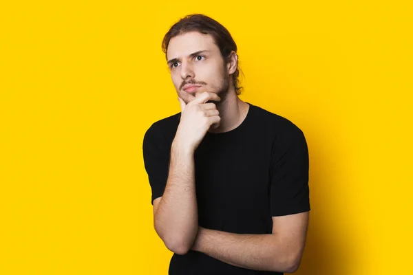 Homem caucasiano pensativo de camisa preta está tocando seu queixo pensando em algo em uma parede de estúdio amarela — Fotografia de Stock