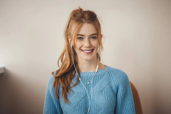 Mujer de jengibre caucásico en un suéter de punto azul está sonriendo a la cámara mientras escucha música usando auriculares en casa — Foto de Stock