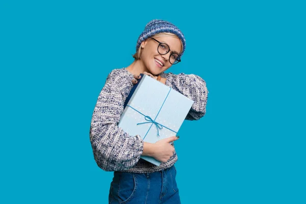 Monokróm kék fénykép egy kaukázusi nőről kalapban és szemüvegben, amint egy ajándékot ölel át a stúdióban, hétköznapi ruhákban mosolyogva a kamerába. — Stock Fotó