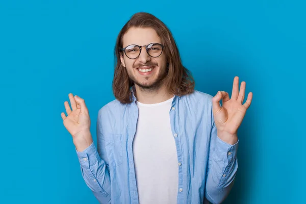 Homem caucasiano com barba e cabelos longos está gesticulando o sinal ok em uma parede de estúdio azul vestindo uma camisa e óculos — Fotografia de Stock