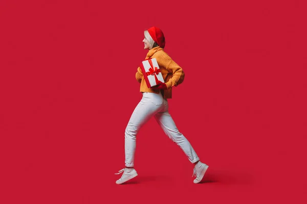Вид збоку фото блондинки з червоним капелюхом, що біжить на студійній стіні з подарунком в руках, посміхаючись під час зимових канікул — стокове фото