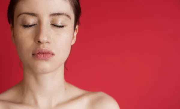 Белая женщина с веснушками позирует с закрытыми глазами и голыми плечами на красной стене студии с свободным пространством — стоковое фото