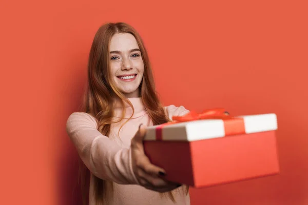 Adorable mujer caucásica con pecas y el pelo rojo está dando a la cámara una caja de regalo sonriendo en una pared de estudio rojo — Foto de Stock