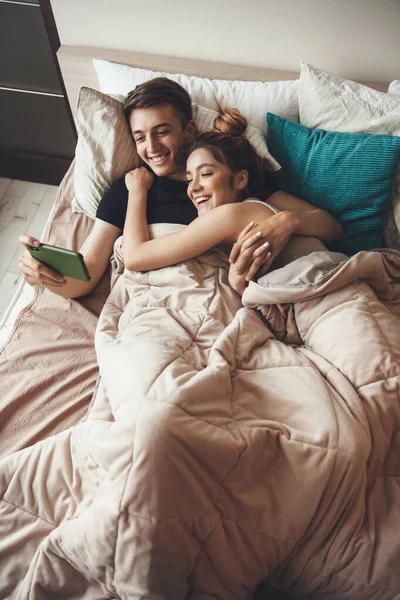 Foto von oben von einem kaukasischen Paar, das im Bett liegt und sich umarmt, während es am Telefon lächelt — Stockfoto