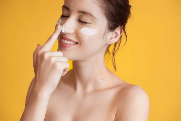 Blanke vrouw met sproeten en naakte schouders is het aanbrengen van een crème op haar wangen en neus terwijl ze glimlacht op een gele studio muur — Stockfoto