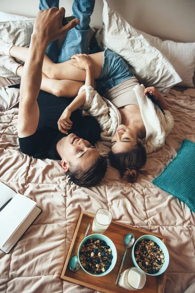 Foto vista superior de um casal caucasiano deitado na cama e fazendo uma selfie enquanto come cereais com leite sorrindo ao telefone — Fotografia de Stock