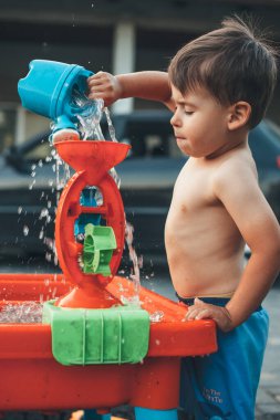 Çıplak göbekli küçük beyaz çocuk arka bahçede su oyuncaklarıyla oynuyor.