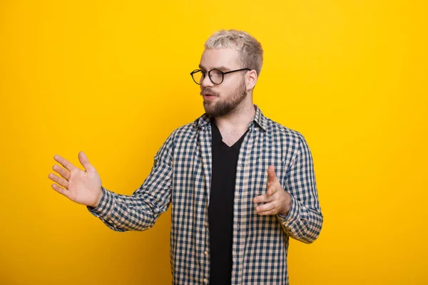 Biały mężczyzna z brodą i blond włosami wyjaśnia coś na żółtej ścianie studia, gestykulując rękami i nosząc okulary. — Zdjęcie stockowe