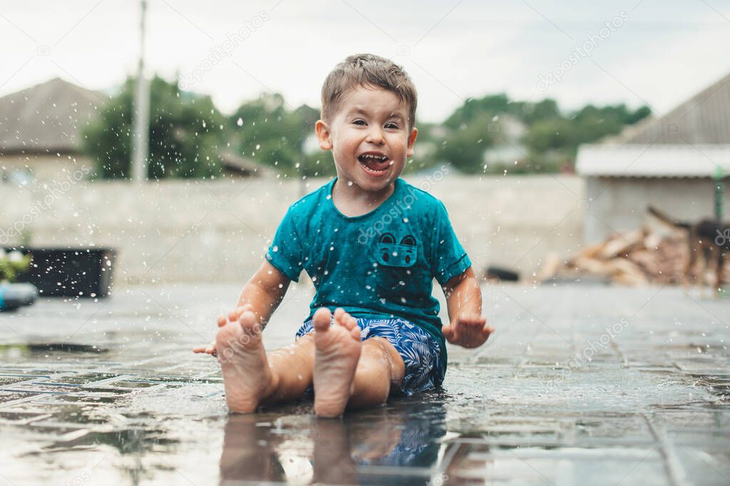 Emotional caucasian boy is playing with water on the ground smiling at camera in the backyard