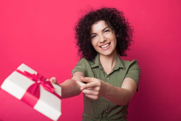Adorable mujer con el pelo rizado está recibiendo un regalo en una pared de estudio rojo sonriendo y sosteniendo una caja de regalo lleno — Foto de Stock