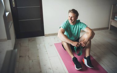 Evde spor kıyafetleri giymiş, sarı saçlı, gülümseyen bir adam.