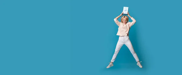 Mujer rubia caucásica está saltando en una pared de estudio azul con espacio libre sosteniendo una caja de regalo en su cabeza y sonrisa — Foto de Stock