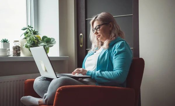 Side view photo of a caucasian senior woman typing something at the laptop while wearing glasses and casual clothes in an armchair — Stock Photo, Image