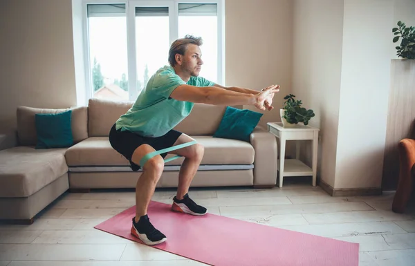 Caucásico hombre está haciendo fitness en casa usando bandas especiales en el suelo mientras se agacha — Foto de Stock