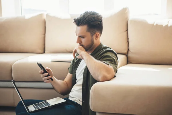Pensativo hombre caucásico está utilizando un teléfono y un ordenador portátil en el suelo cerca de un sofá mientras trabaja de forma remota —  Fotos de Stock
