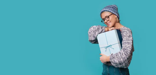 Kaukasische Frau mit Hut und Brille umarmt lächelnd ein Geschenk an einer blauen Studiowand mit freiem Raum — Stockfoto