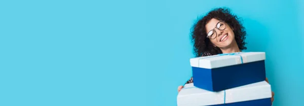 Caucásico rizado dama de pelo rizado está sonriendo a la cámara con gafas mientras sostiene algunas cajas de regalo en una pared de estudio azul con espacio libre — Foto de Stock