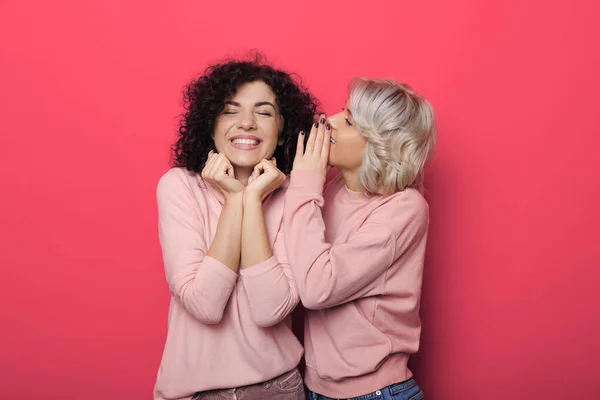 Adorable woman with blonde hair is whispering something to her brunette curly haired sister on red studio wall — Stock Photo, Image