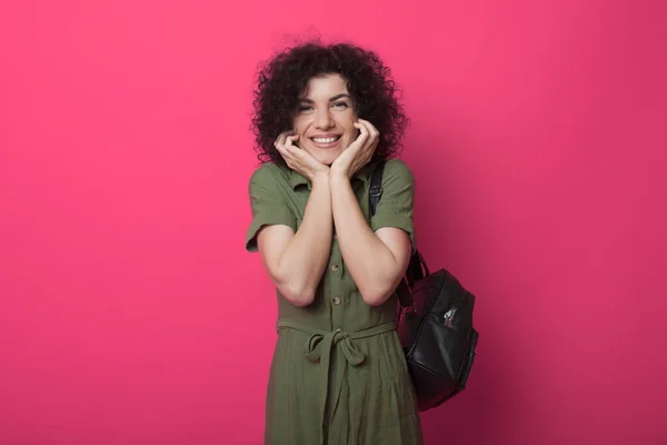 Adorabile studente dai capelli ricci sta sorridendo e guardando la fotocamera che tiene una borsa su una parete rossa dello studio — Foto Stock